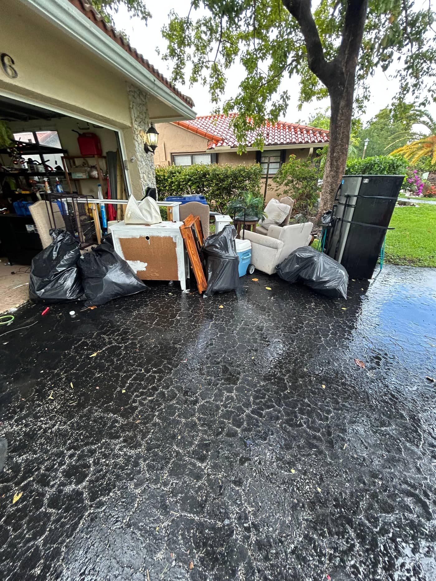 A driveway with black bags, an armchair, a speaker cabinet and boxes set out in front of an open garage