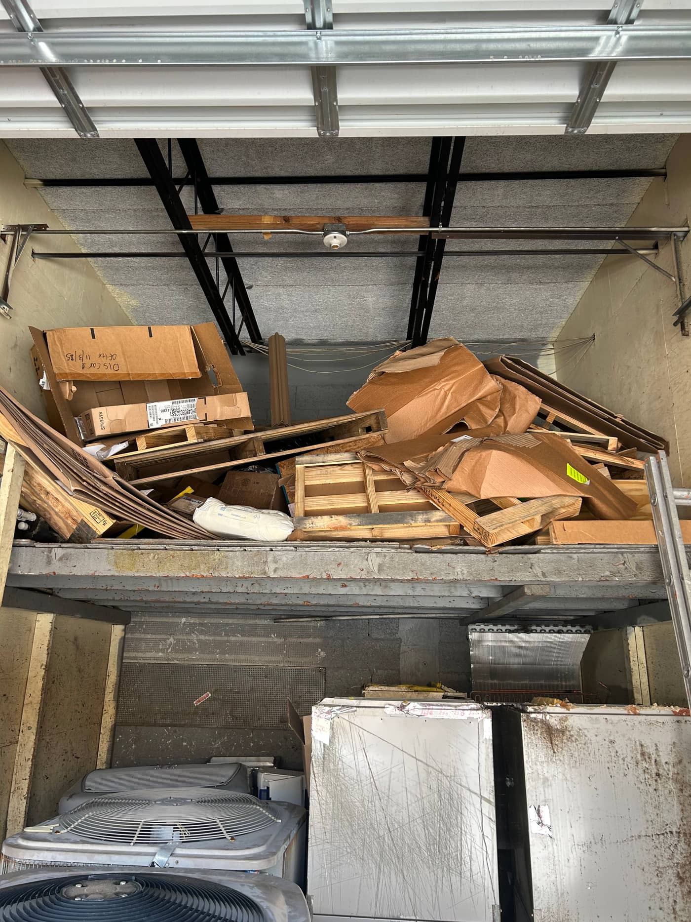 A garage with offcut lumber, plywood and flattened cardboard piled across the whole floor, in front of an air handler and stacked panels