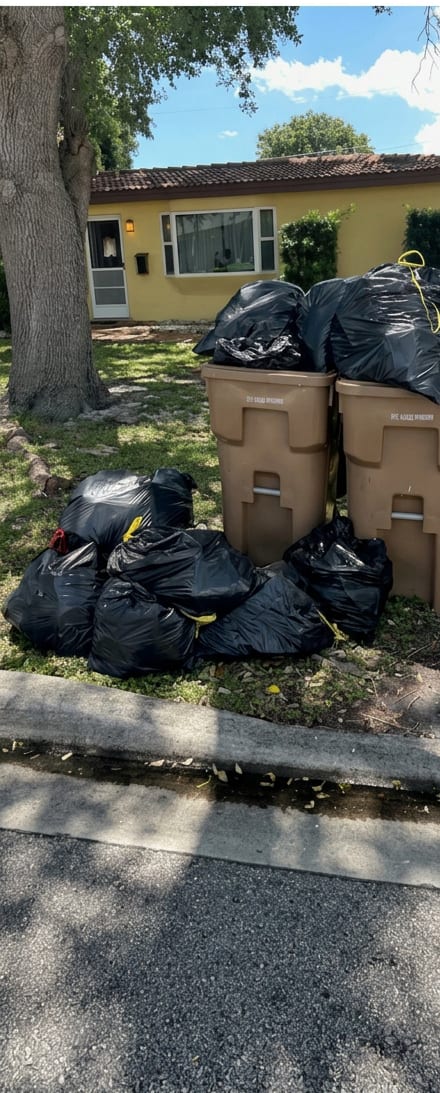 A dozen black trash bags piled around two overflowing city bins on the front lawn of a yellow South Florida house