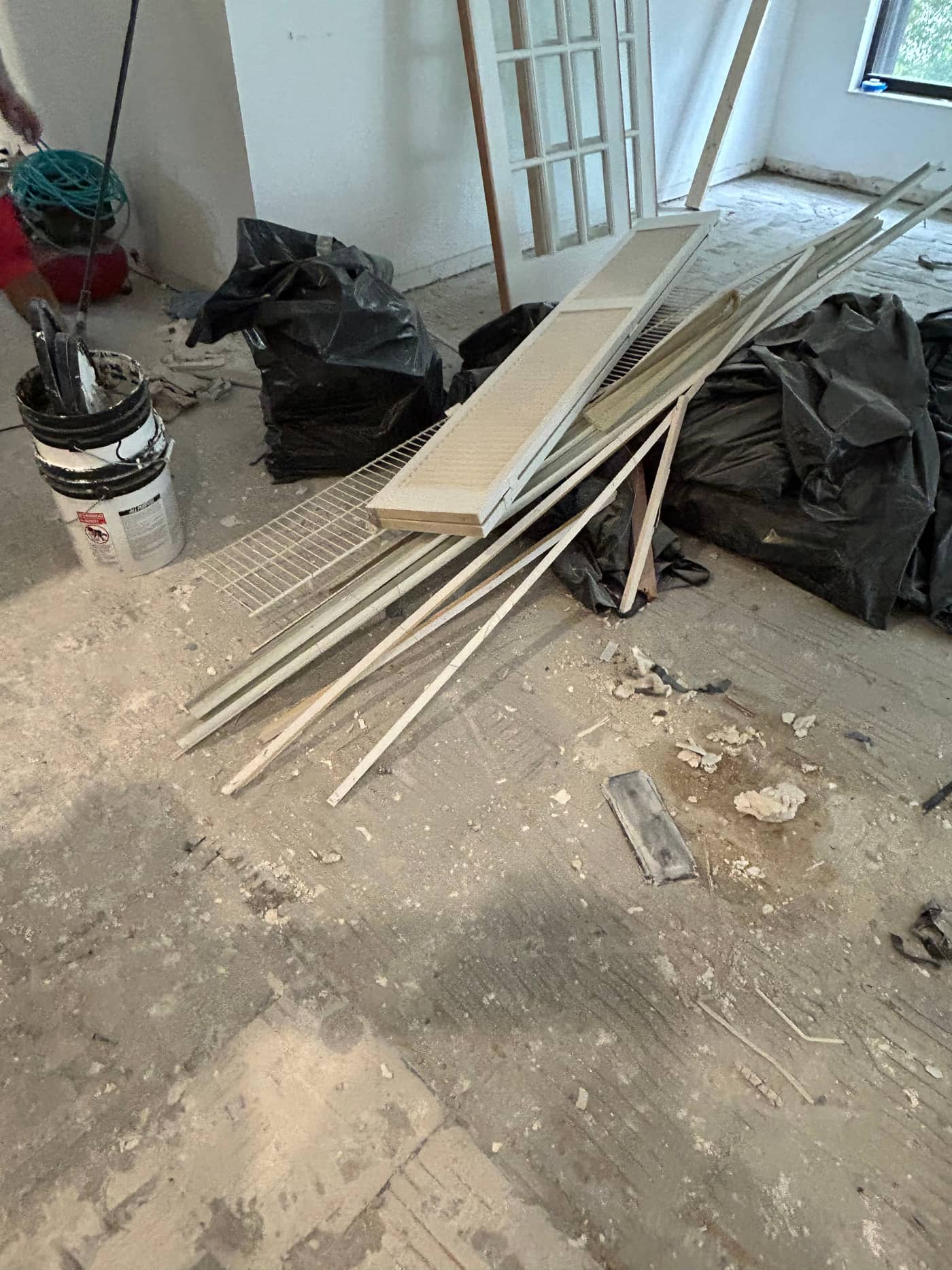 A room mid-renovation with black bags, stacked lengths of trim and moulding, a bucket and loose debris on a bare floor