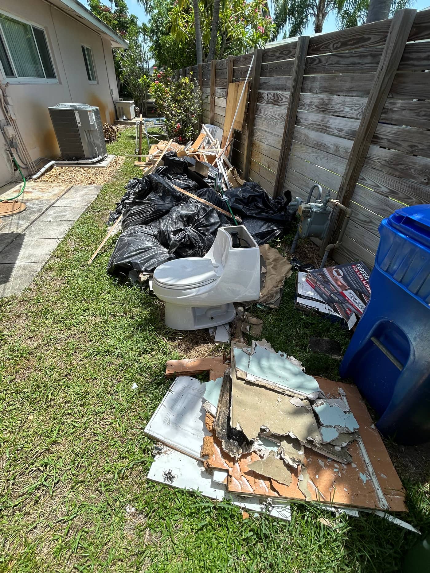 A side yard with a toilet, black bags, broken board and tile piled along a wooden fence beside a recycling bin