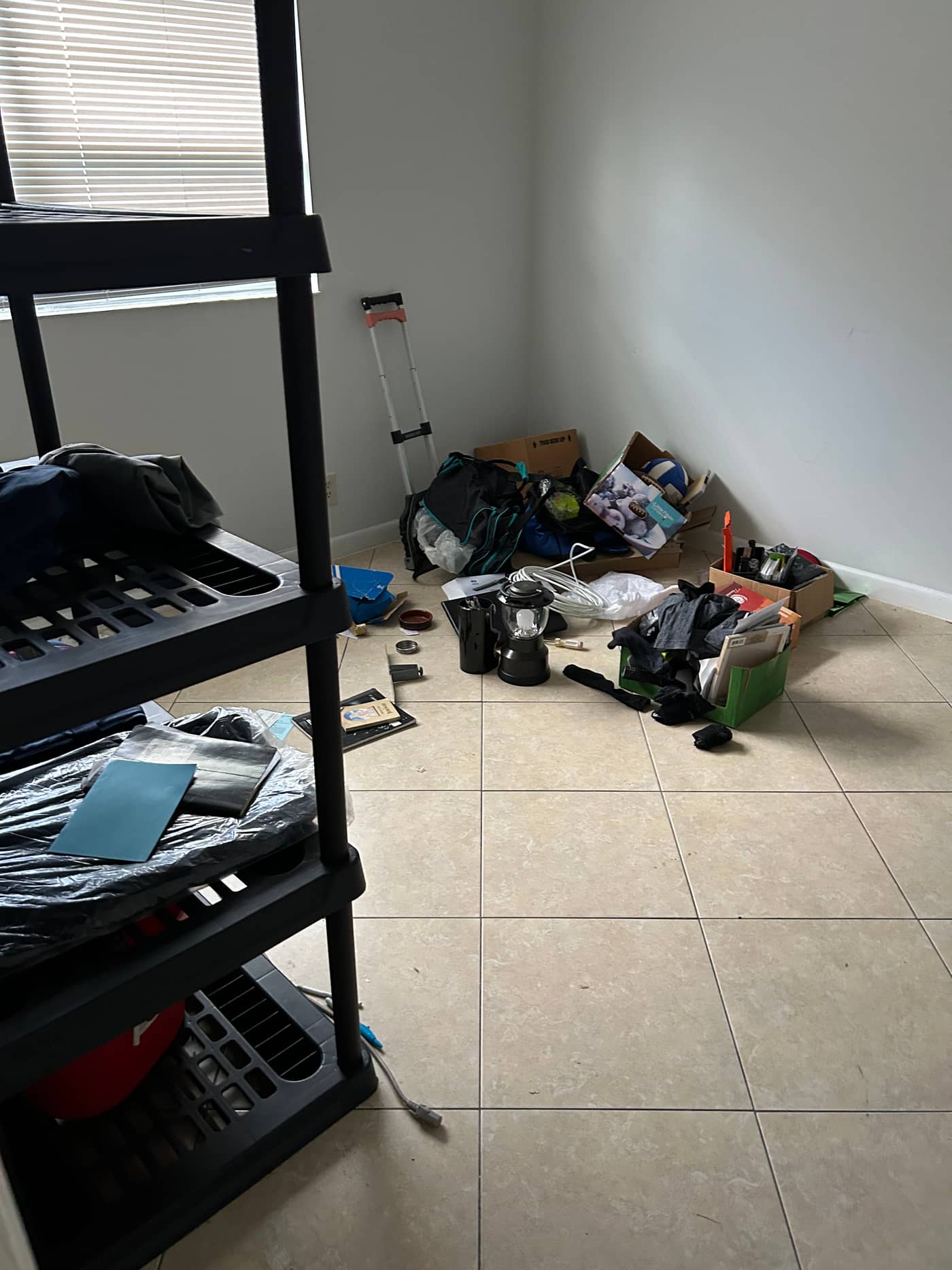 A tiled room with a black shelving unit, a step ladder, bags of clothing and loose household items piled into the corner
