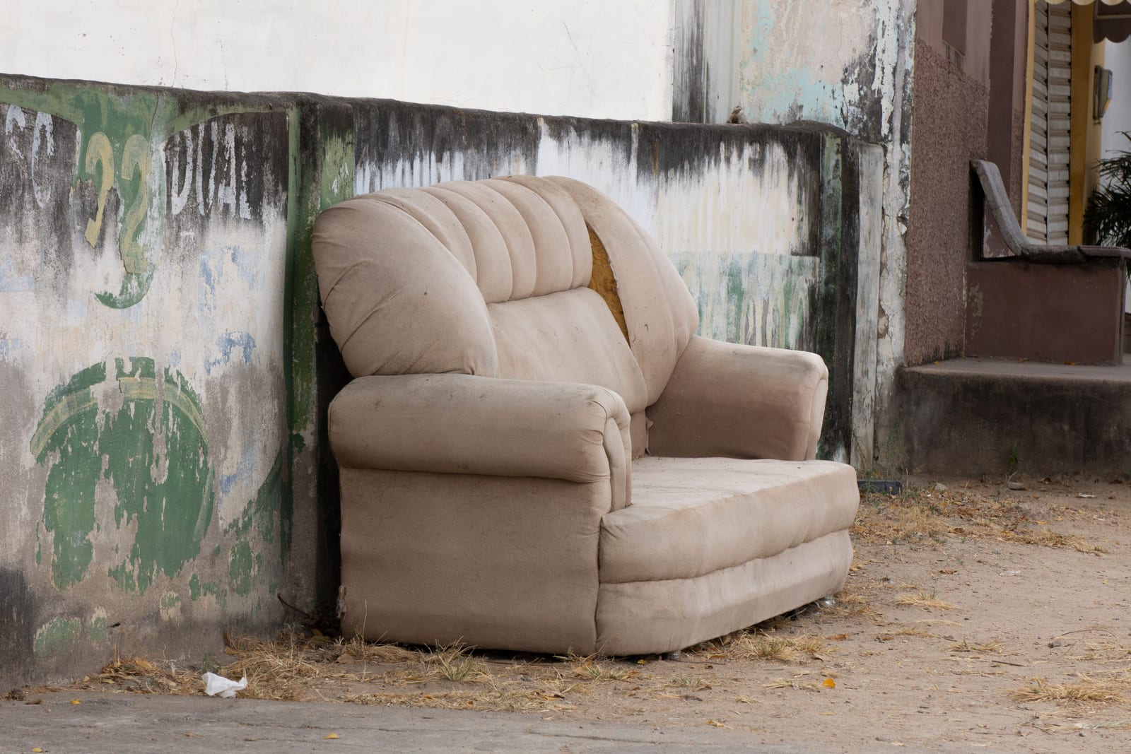 A worn armchair left out on the kerb against a bare wall, waiting for a pickup that may not take it