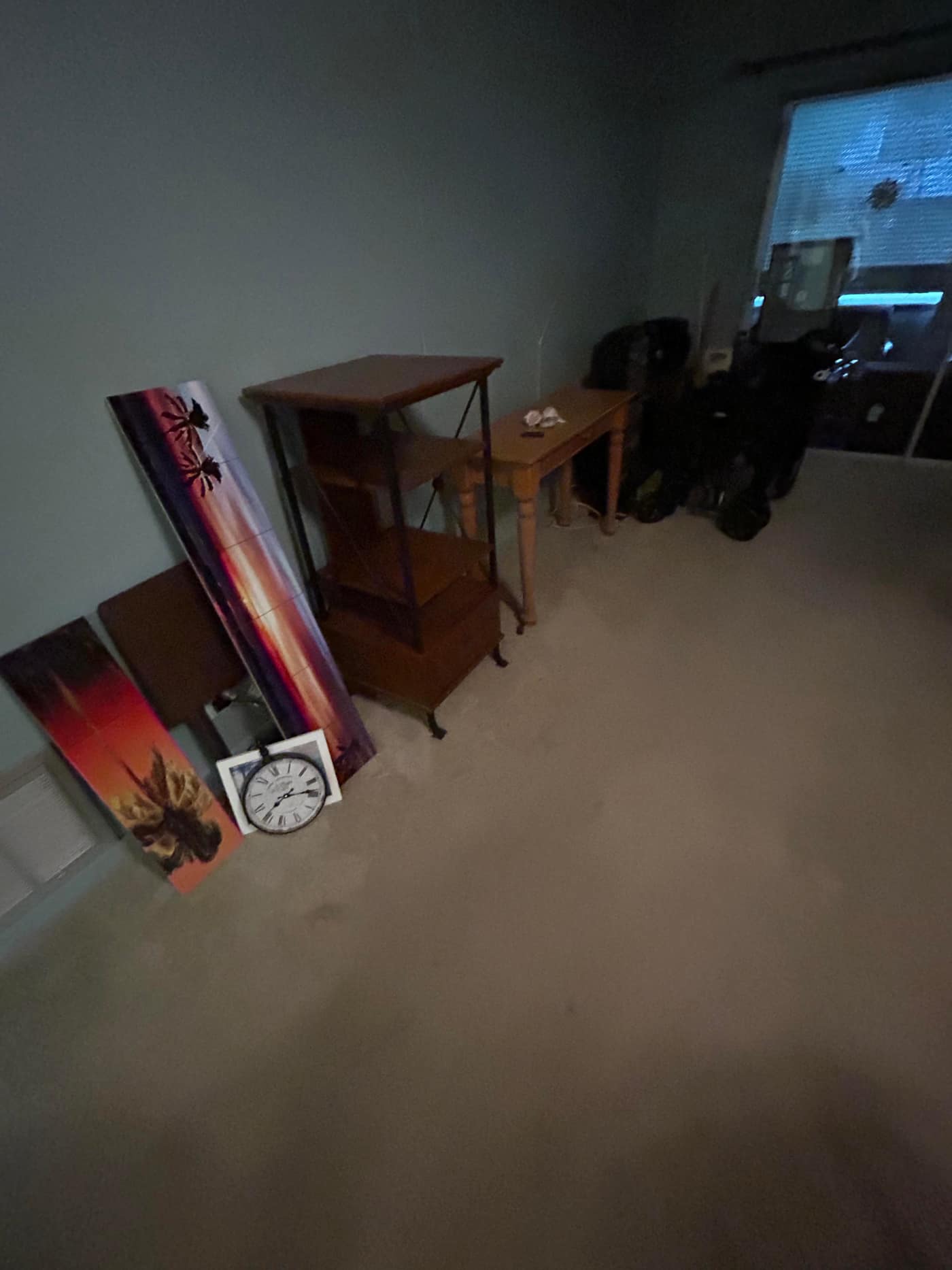 A dim carpeted apartment room with framed sunset canvases and a wall clock leaning against the wall, two wooden side tables and a shelving unit beside them, and bagged belongings piled in the far corner