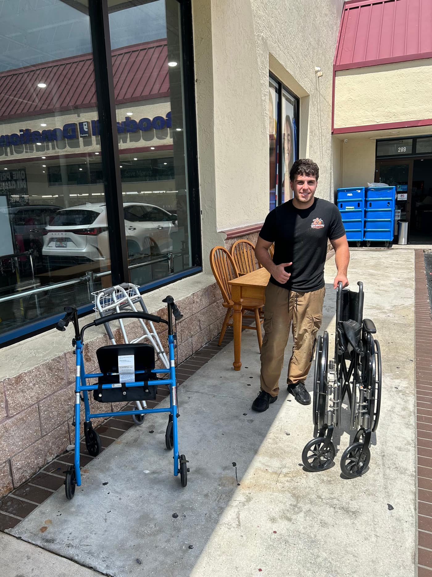 Johnny outside a Goodwill donation door with a folded wheelchair, two rollators and a wooden dining table and chairs off the truck