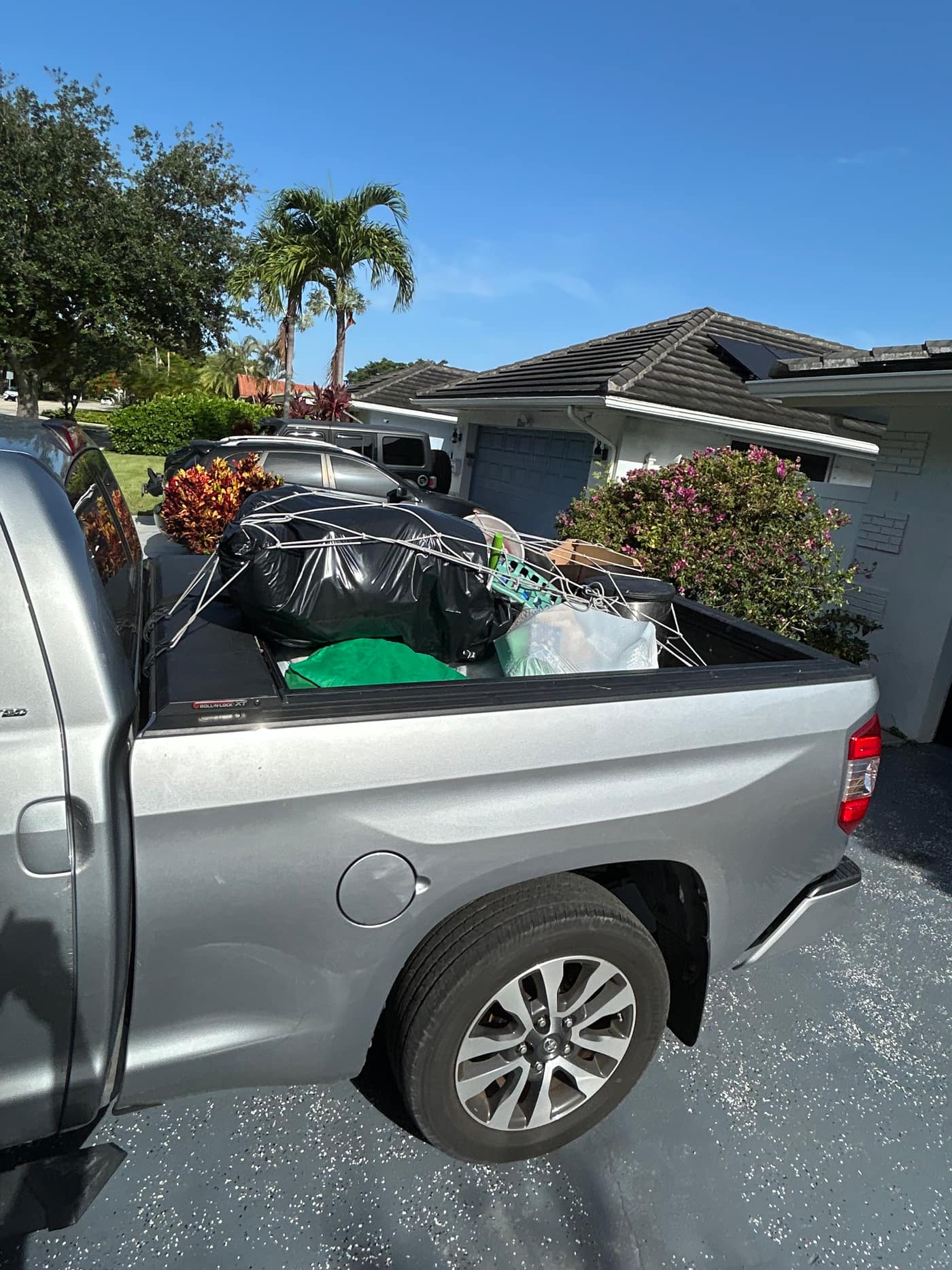 The Gym Junksters pickup on a South Florida driveway with its bed loaded. A wrapped black bundle, a green tarp, a broom, cardboard and a bin, strapped down under a cargo net.